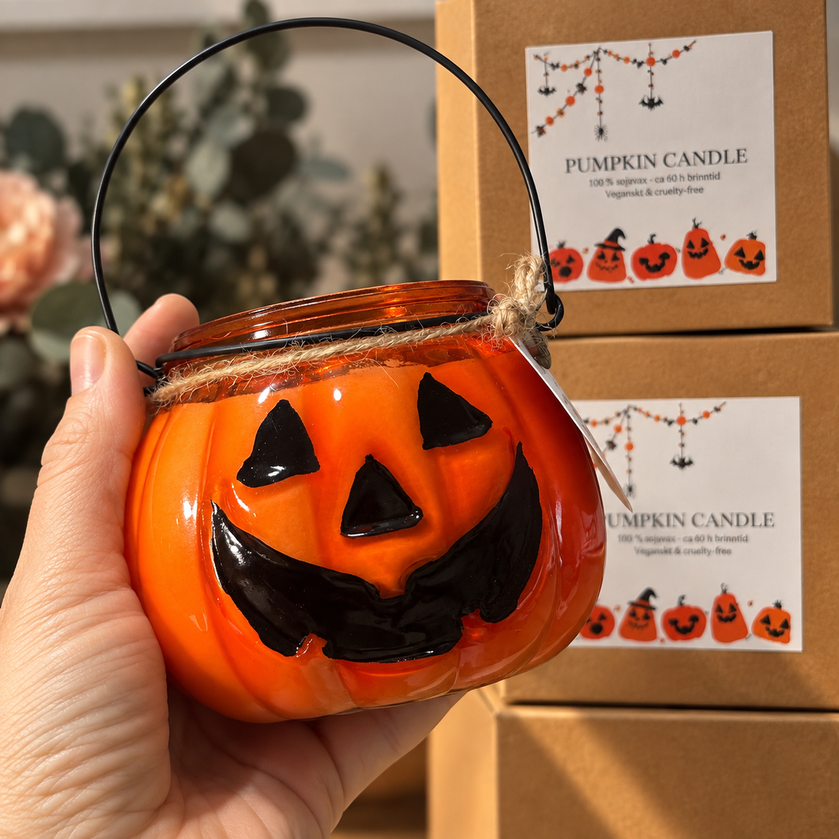 Närbild på Pumpkin Glow, ett handgjort Halloween-doftljus i orange pumpaformad glaslykta med svart handtag, som hålls i handen framför presentförpackningar.